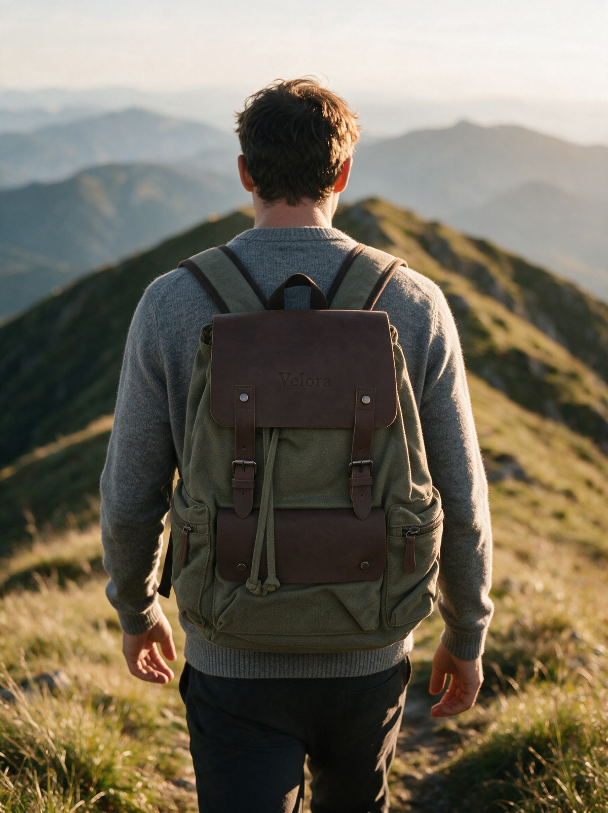 Mann trägt den olivgrünen Canvas Rucksack mit Kunstleder-Klappe auf einem Bergpfad im Abendlicht, Blick über die Bergkette